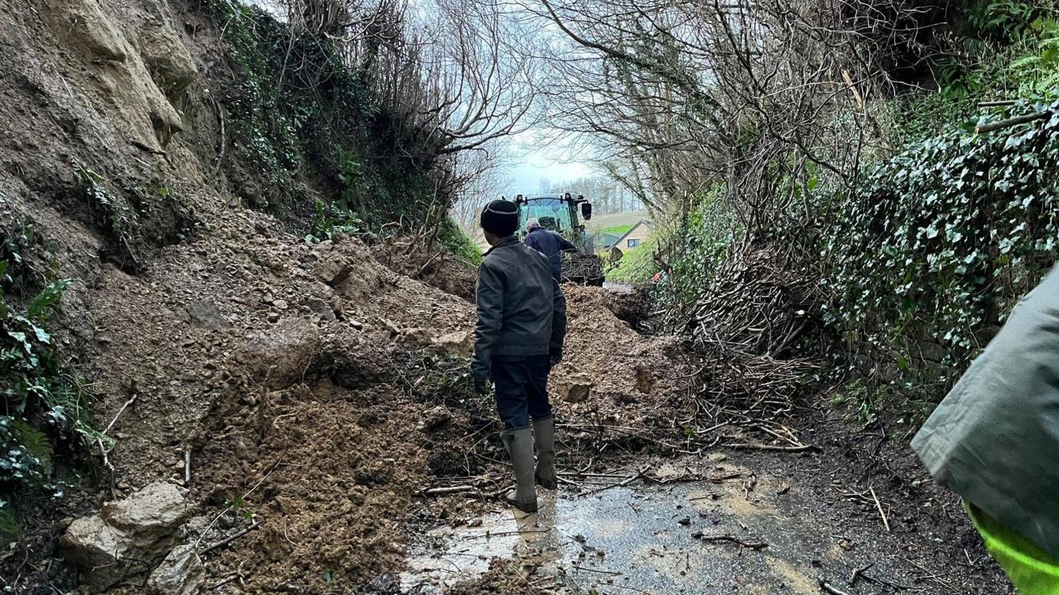 A landslip in Barrington over a small road. A large amount of rocks and debris are seen piled up and in the distance a tractor is visible as a small group of people try to clear the road. Each side of the road there is a steep green verge