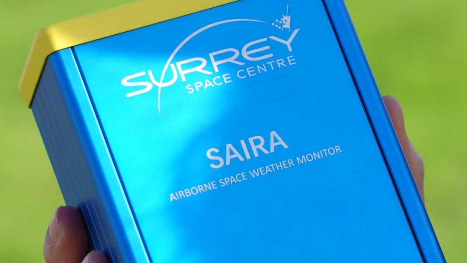 A blue box has the words Surrey Space Centre Saira Airborne Space Weather Monitor on it. It is held on someone's hands, to give you an idea of its size.