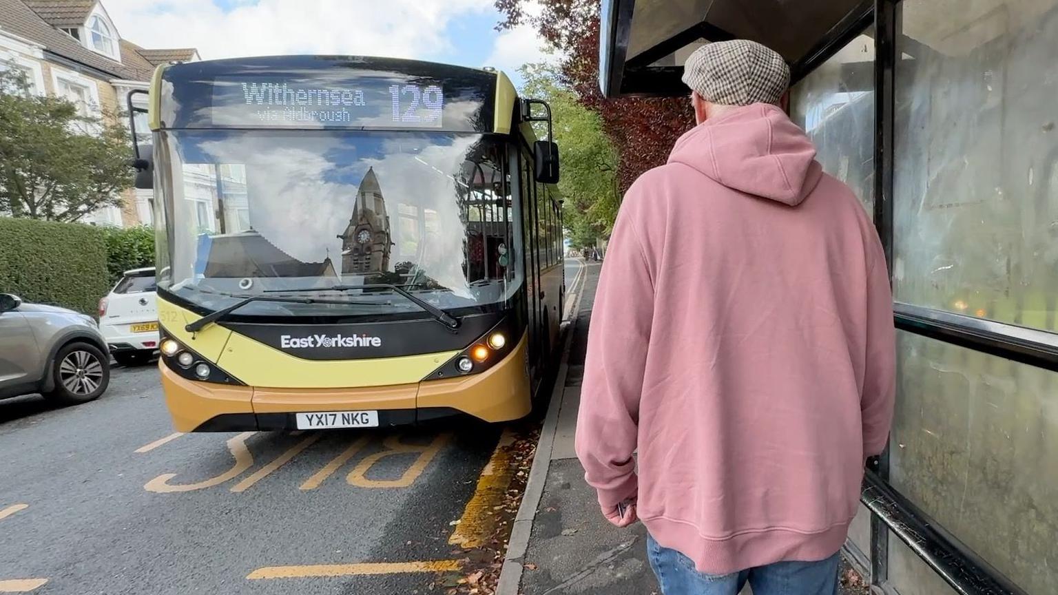 A man in a pink hoodie, jeans and a checked flat cap prepares to board a single decker bus at a bus stop. The bus display says it is the 129 to Withernsea. The bus is yellow and black, and Ian has his back to the camera.