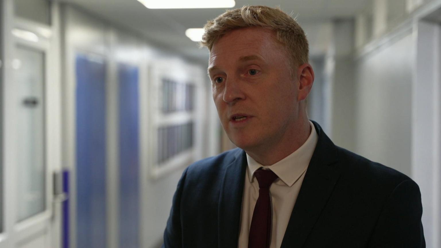 Gareth Lee has short hair and is wearing a dark suit with a white shirt and red tie.