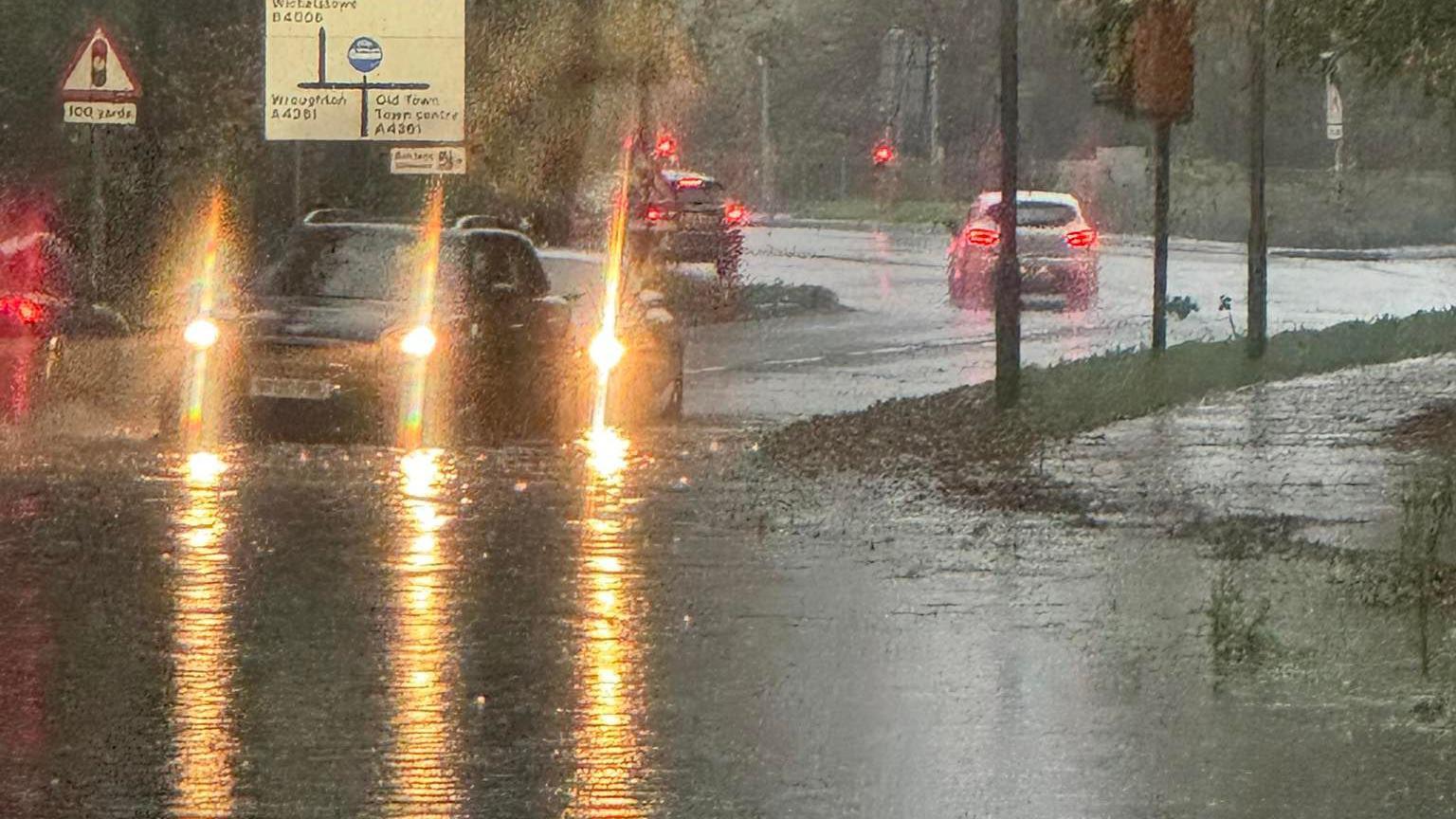 Cars turning round a corner where there heavy rainfall has created a large puddle. The water has flooded the pavement and road. 