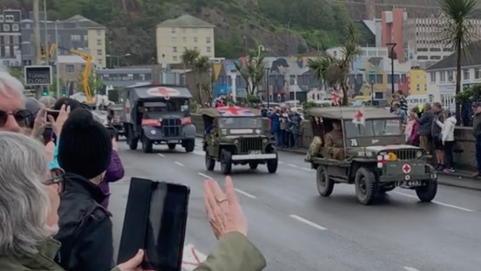 A picture of a lady waving at the cars. The cars are small and green in colour with people stood either side of the road.