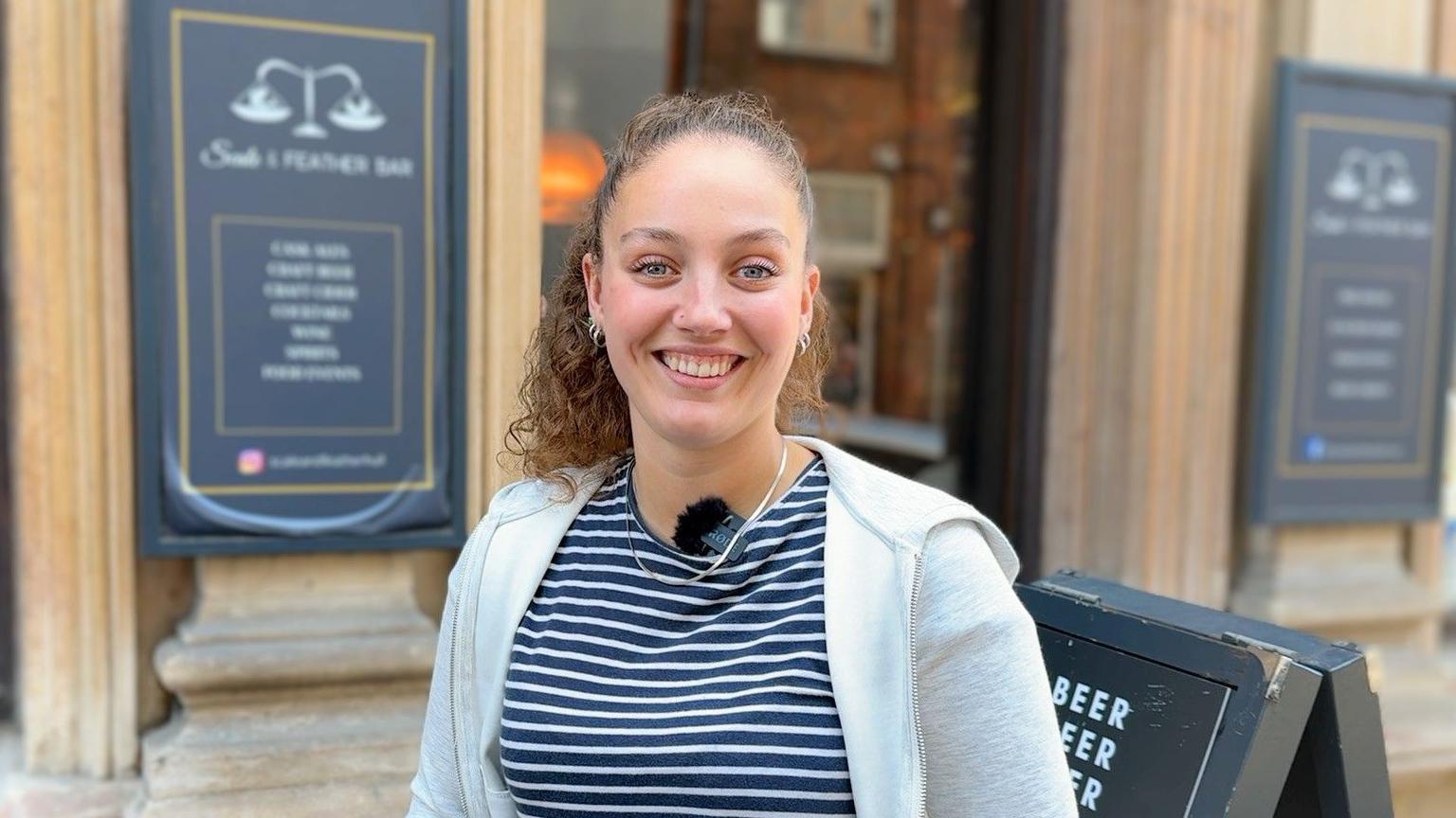 Alex looks into the lens smiling. She has curly brown hair tied back in a ponytail and is wearing a navy and white striped t-shirt with a grey hoodie.