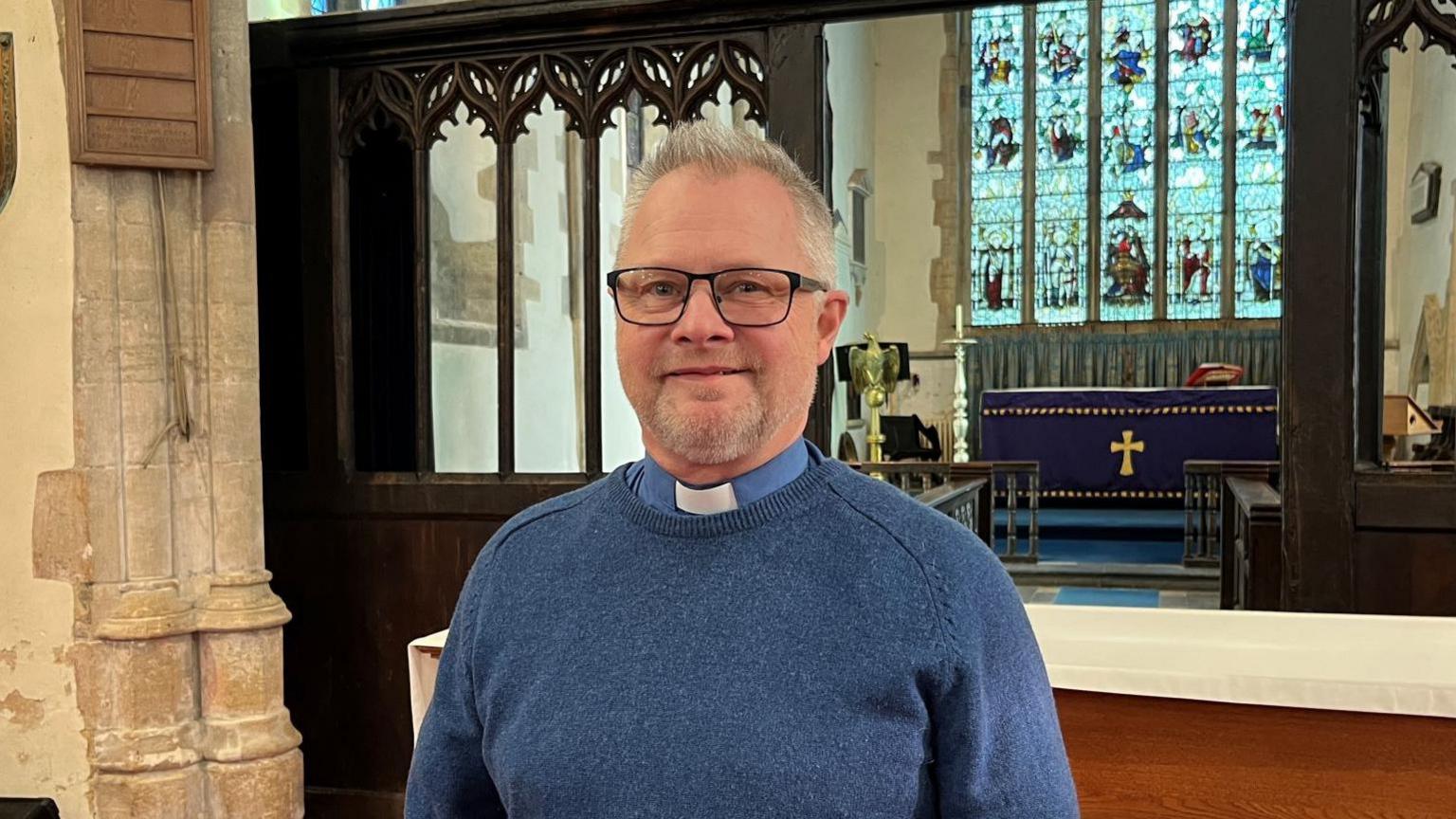 A man wearing a blue jumper and glasses. He has a blue and white dog collar. He is stood inside a church with stained glass windows behind.