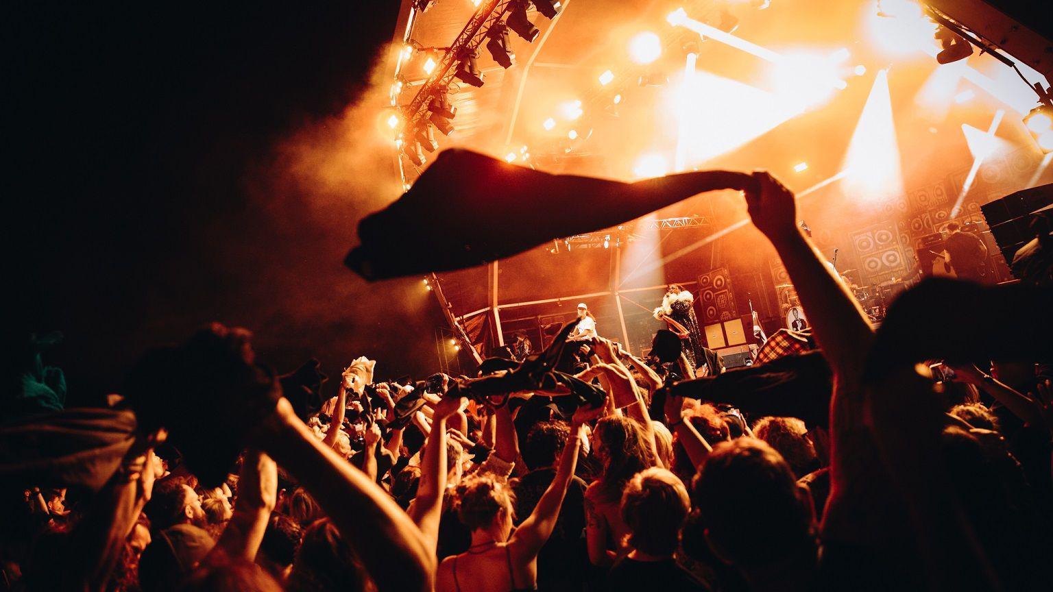 Rock band Skindred perform on stage at Bristol Sounds. The band and the audience are illuminated by floodlights which are casting an orange glow. The picture is taken from within the audience, looking up at the stage