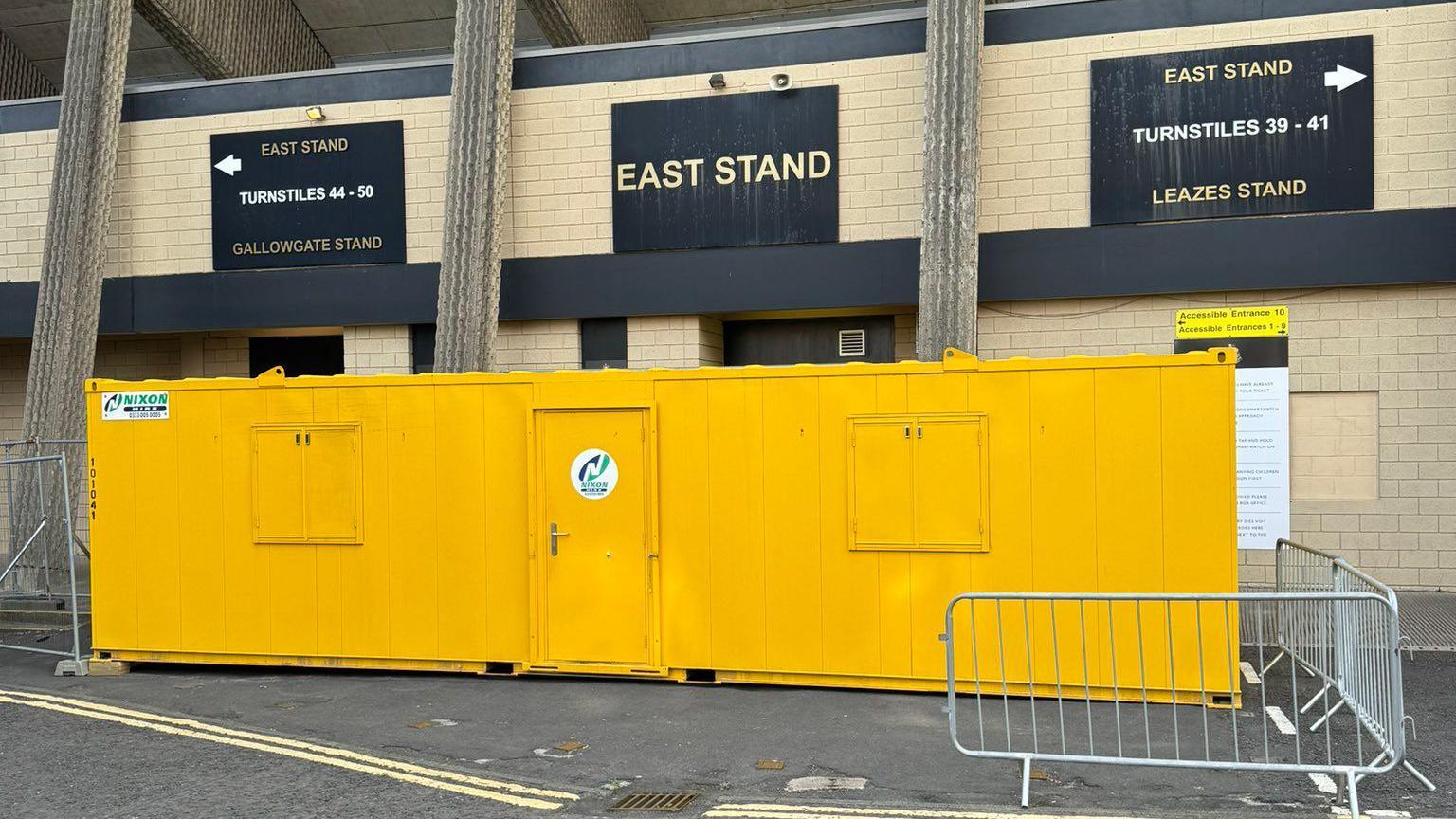 A yellow portable building placed on the land outside of St James' Park. It has a door in the centre of the building.  A small amount of metal fencing has been put up around the land.