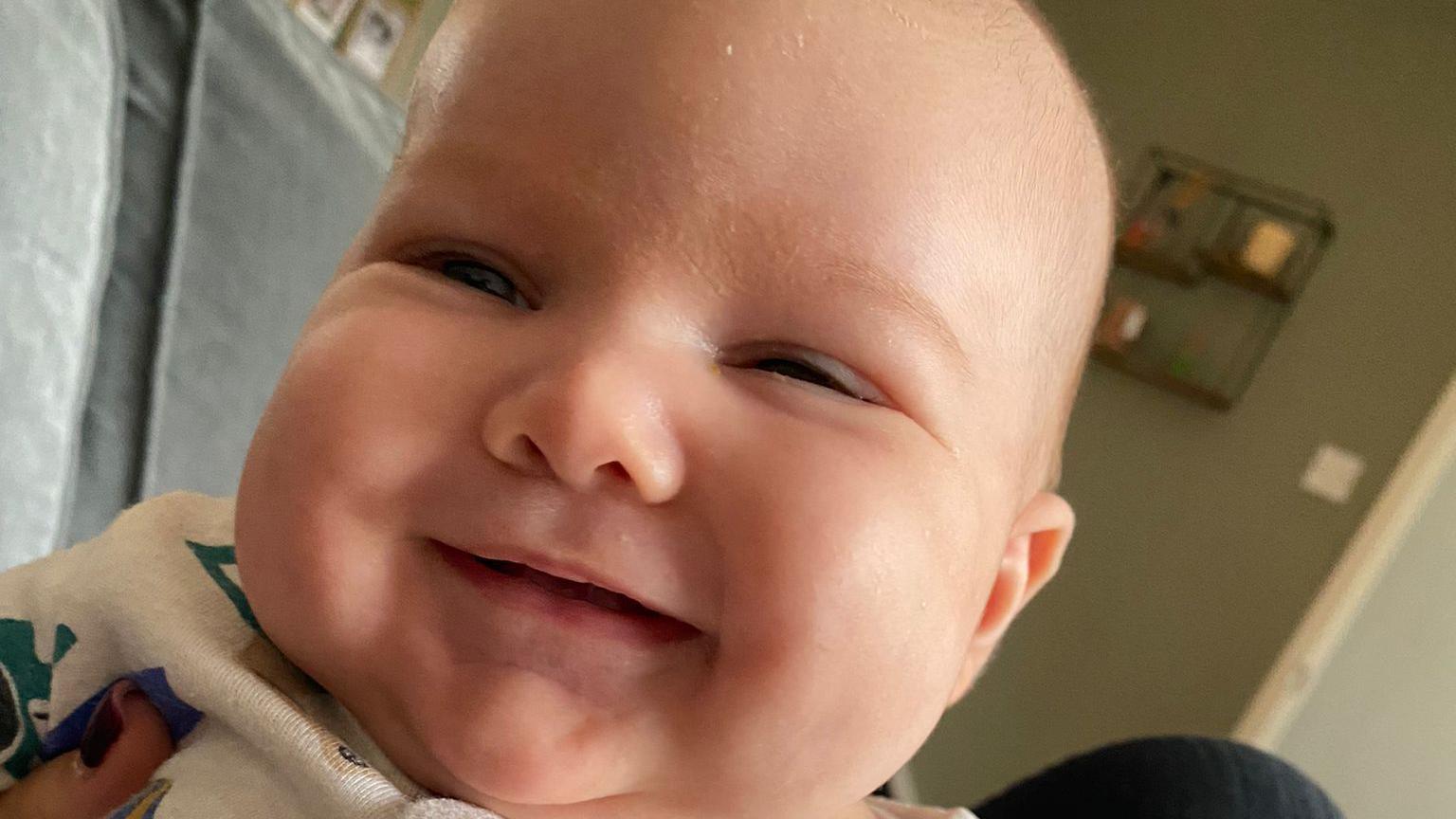 A baby boy smiles at the camera in a close up picture. He is wearing a white baby grow while an adult holds him.