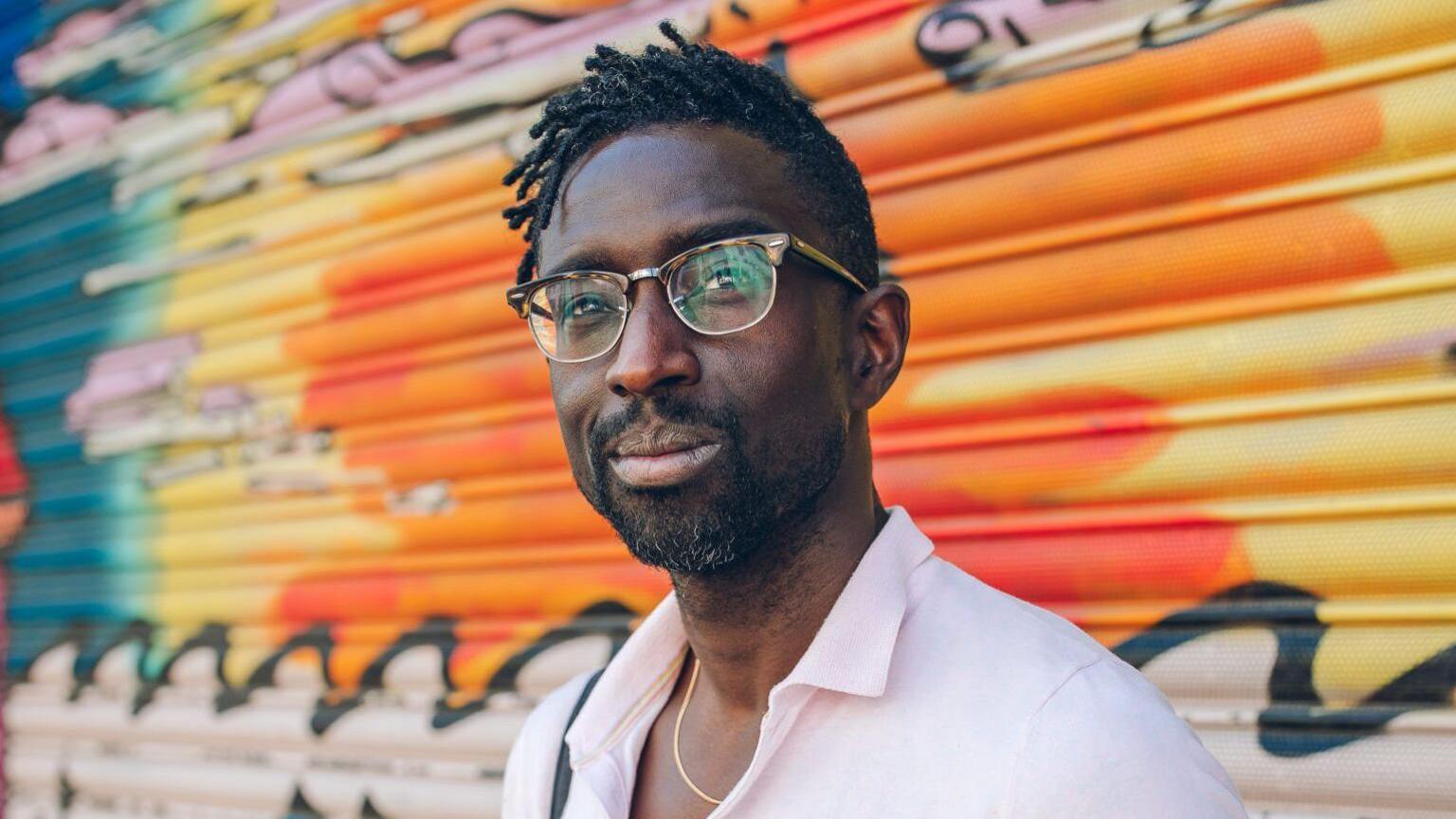 Jeffrey Boakye, wearing an open buttoned white shirt standing in front of a garage door painted in brightly-coloured graffiti 