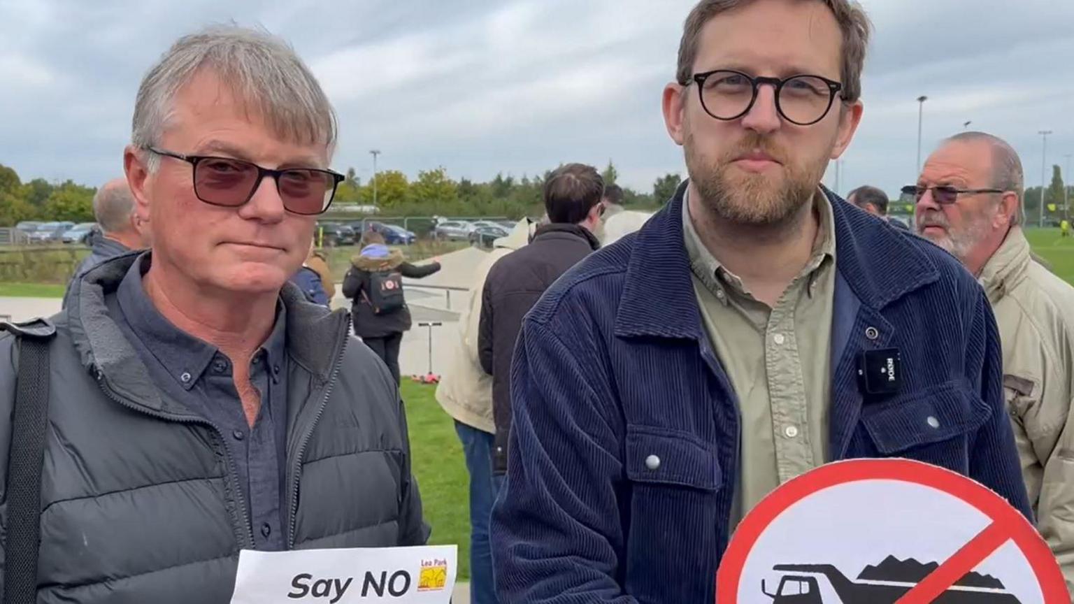 Henley and Thame MP Freddie Van Mierlo attending the demonstration, holding a sign that had a lorry crossed out. Another resident is next to him, holding a sheet of paper that says "Says NO". Other people could be seen behind them. It's an overcast day.