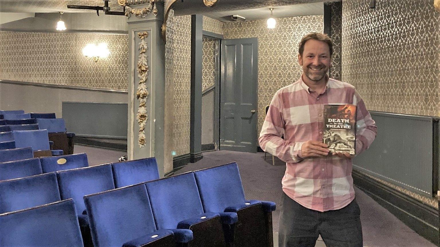 Chris Wood wearing a pink and white plaid shirt and blue-grey jeans. He is standing in a theatre holding his book, Death in the Theatre, and smiling at the camera. The theatre has rows of blue velvet seats and ornate wallpaper and decorative molding.