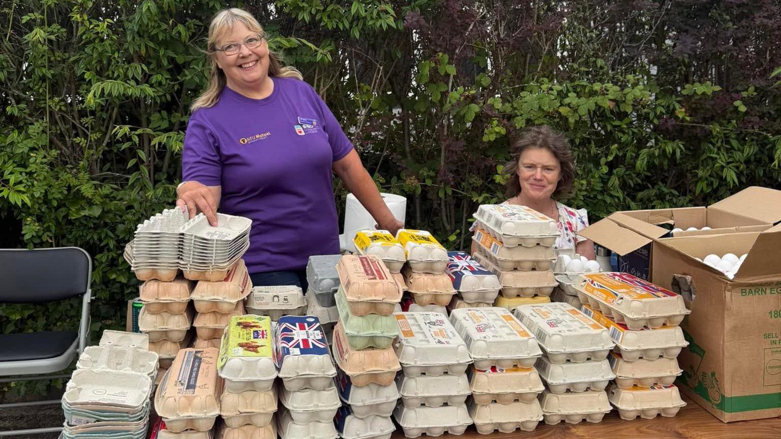 Two women are behind a table with a large collection of egg boxes on it. The woman on the right is sitting down. The woman on the left is standing up and wearing a purple T-shirt. A chair is to the left of the table and a cardboard box with white eggs in it, to the right. There are trees in the background. 