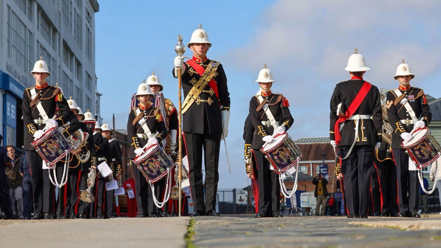 A military band stands in the grounds of Portsmouth Historic Dockyard, they are wearing ceremonial uniform with red stripes down the sides of the trousers and white peaked hats, standing in rows. The dockyard warehouse can be seen in the background.