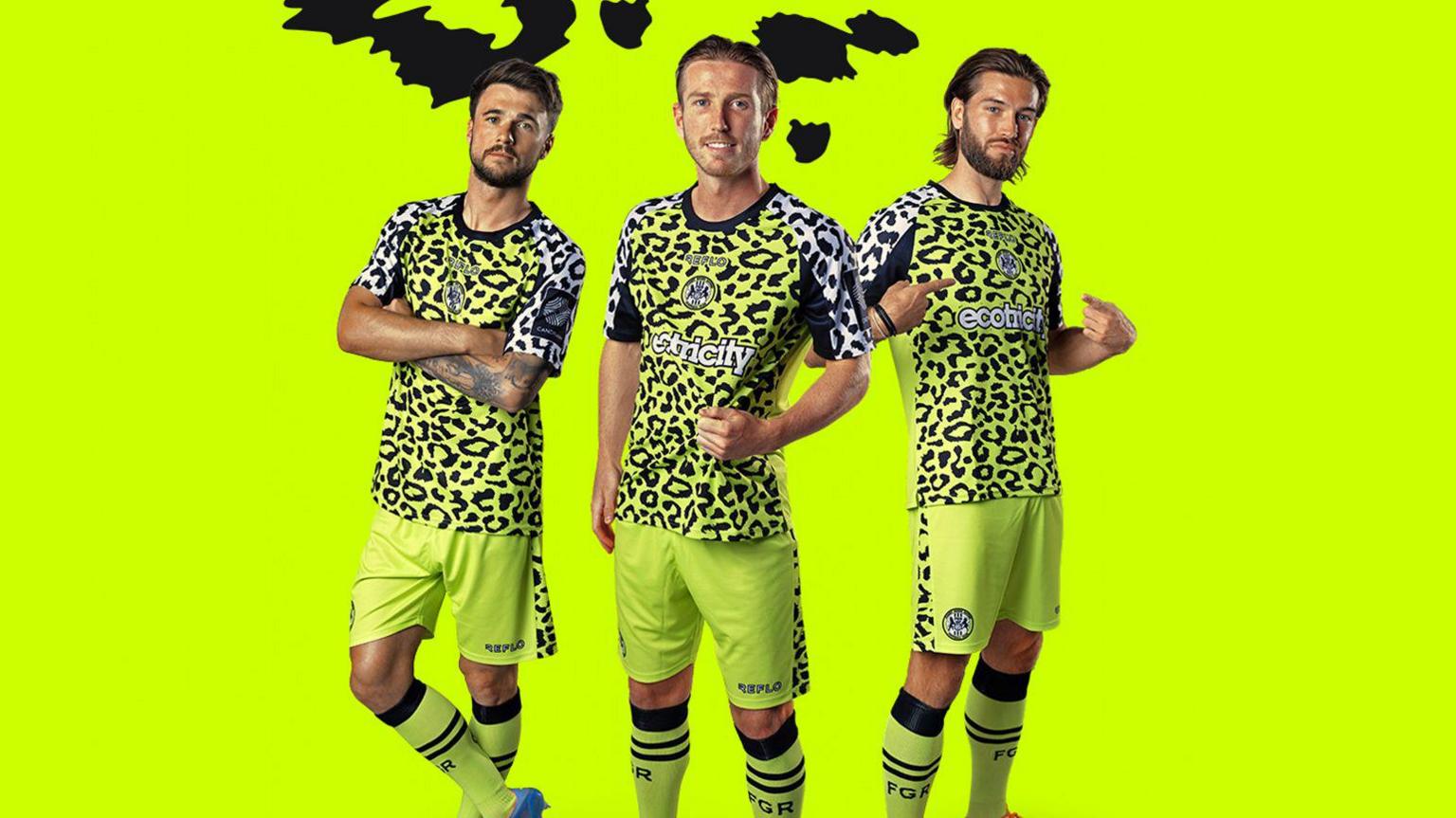 three footballers stand together wearing a fluorescent green kit with the top covered in black spots similar to the markings of a leopard.