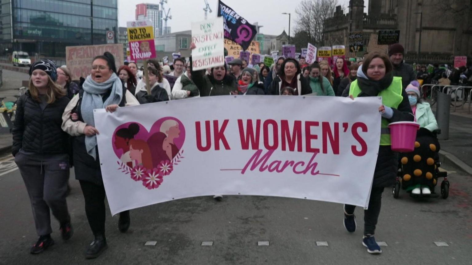 Women hold a large white and pink banner saying UK Women's March while marching near Manchester Cathedral