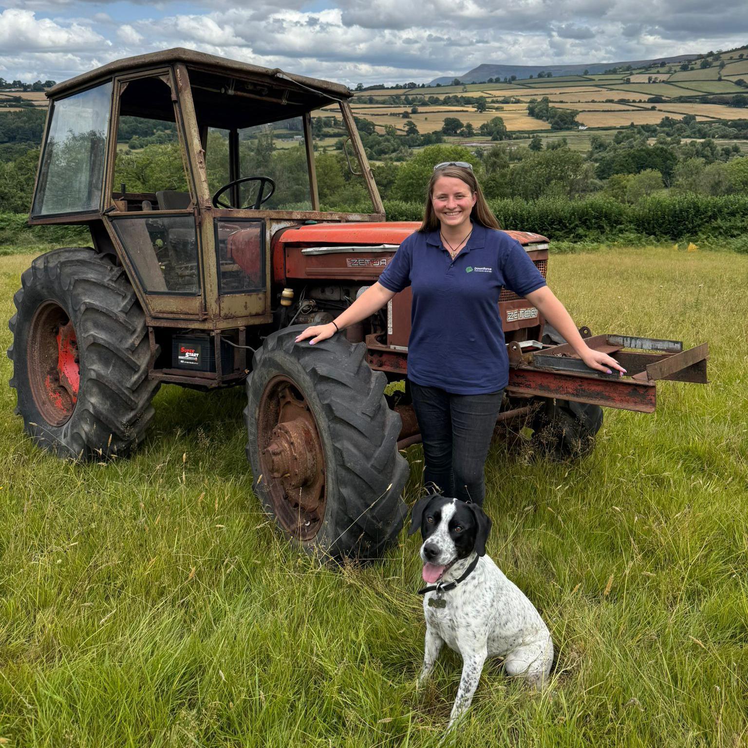 Liz Tree wearing a blue T-shirt and black trousers standing in front of an old tractor in a field with a black and white dog sitting in front of her. More fields and hills can be seen behind her in the background