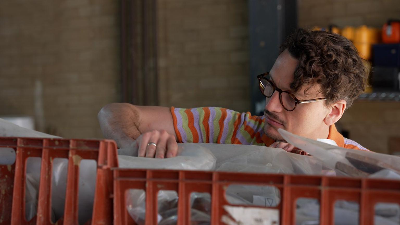A candid shot of Tom Booth, who wears glasses and a colourful stripey t-shirt, looking through a box of archaeological finds.