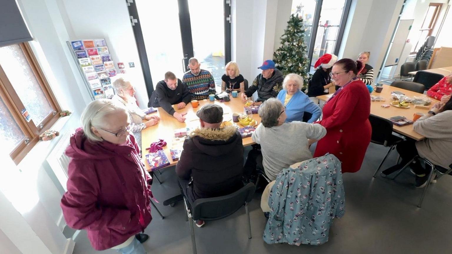 Men and women sit around two tables at Middleport Matters. One woman in purple and another in red are standing up.