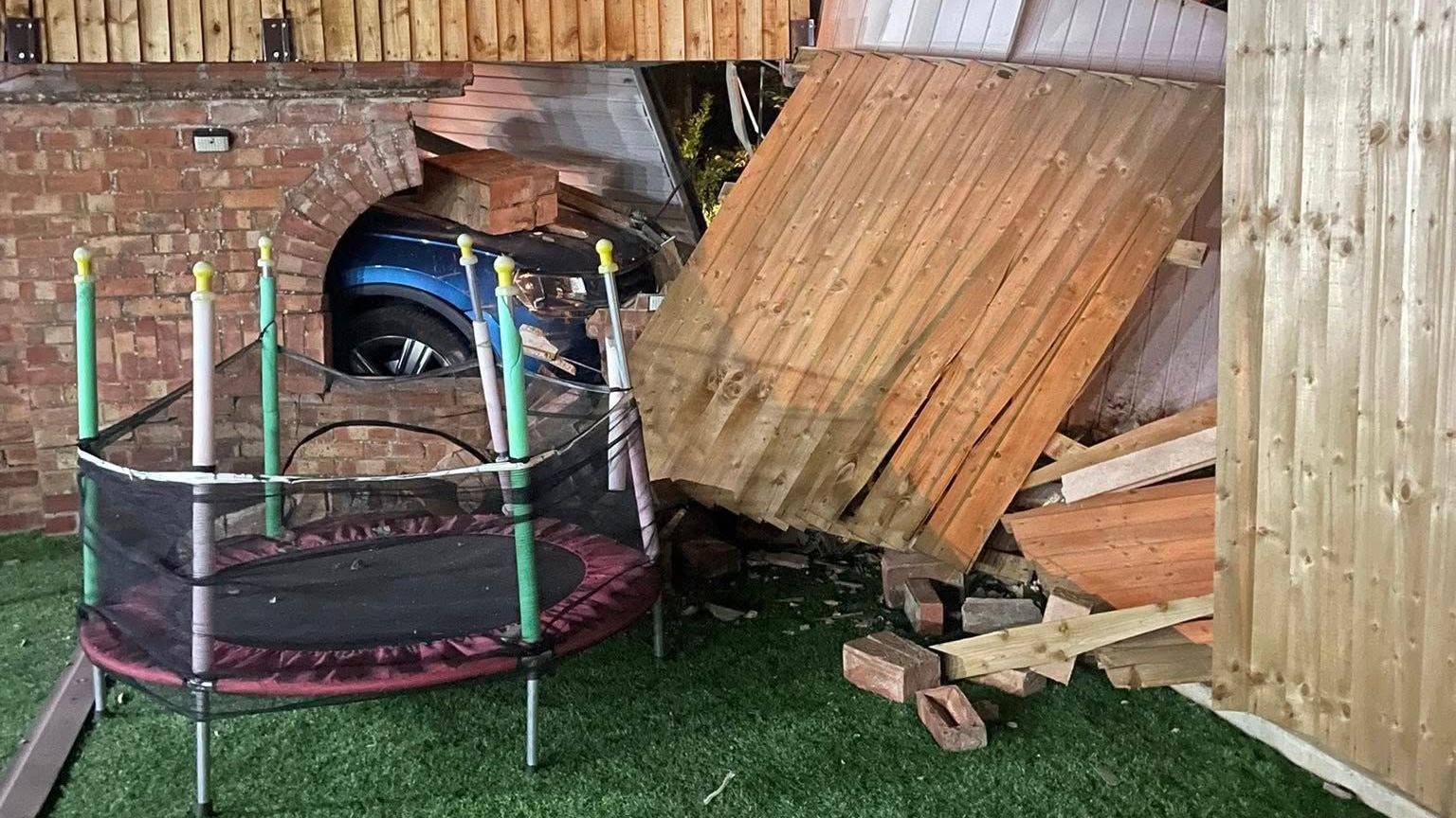 Part of a blue car after it has been pushed into brickwork in a garden after another vehicle crashed behind it. Bricks are strewn on a lawn and fence panels are broken near a child's trampoline.