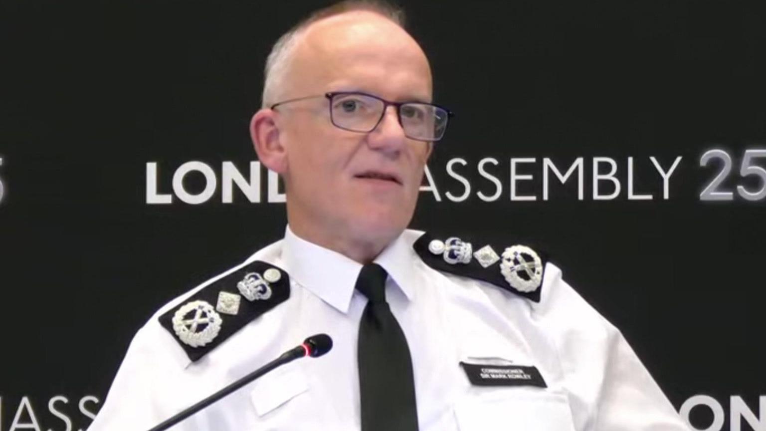 Sir Mark Rowley is seen sitting at a table speaking into a microphone, with a London Assembly backdrop behind him.
