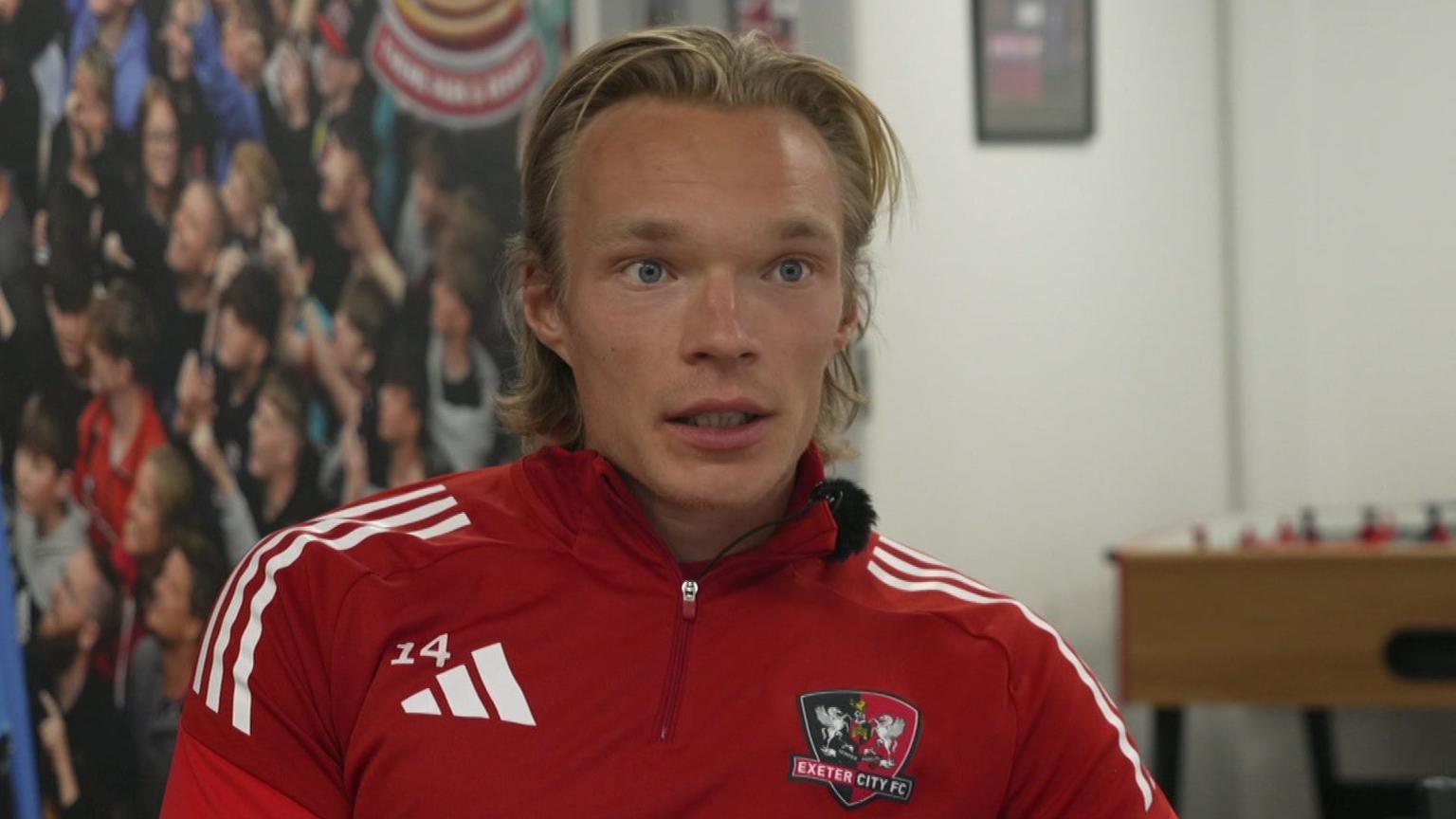 A blond haired football player dressed in an Exeter City Football club red tracksuit.