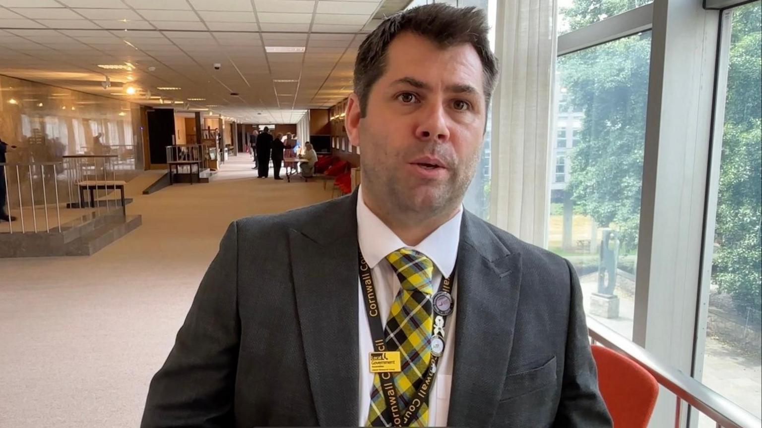 Leigh Frost, a middle-aged man with short dark hair and light stubble, in a suit with a yellow, black and blue Cornish tartan tie.