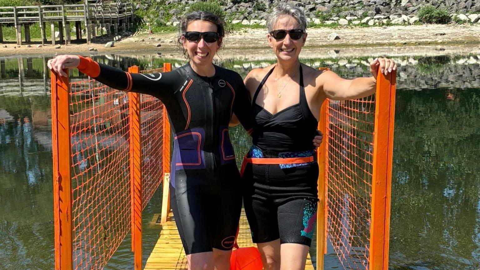 Two women wearing sunglasses are standing on a wooden ramp with orange fencing on the sides. They are standing next to each other, both with an arm around the other, and holding onto the fence with the other arm. The lake is behind them and they are wearing swimming suits.