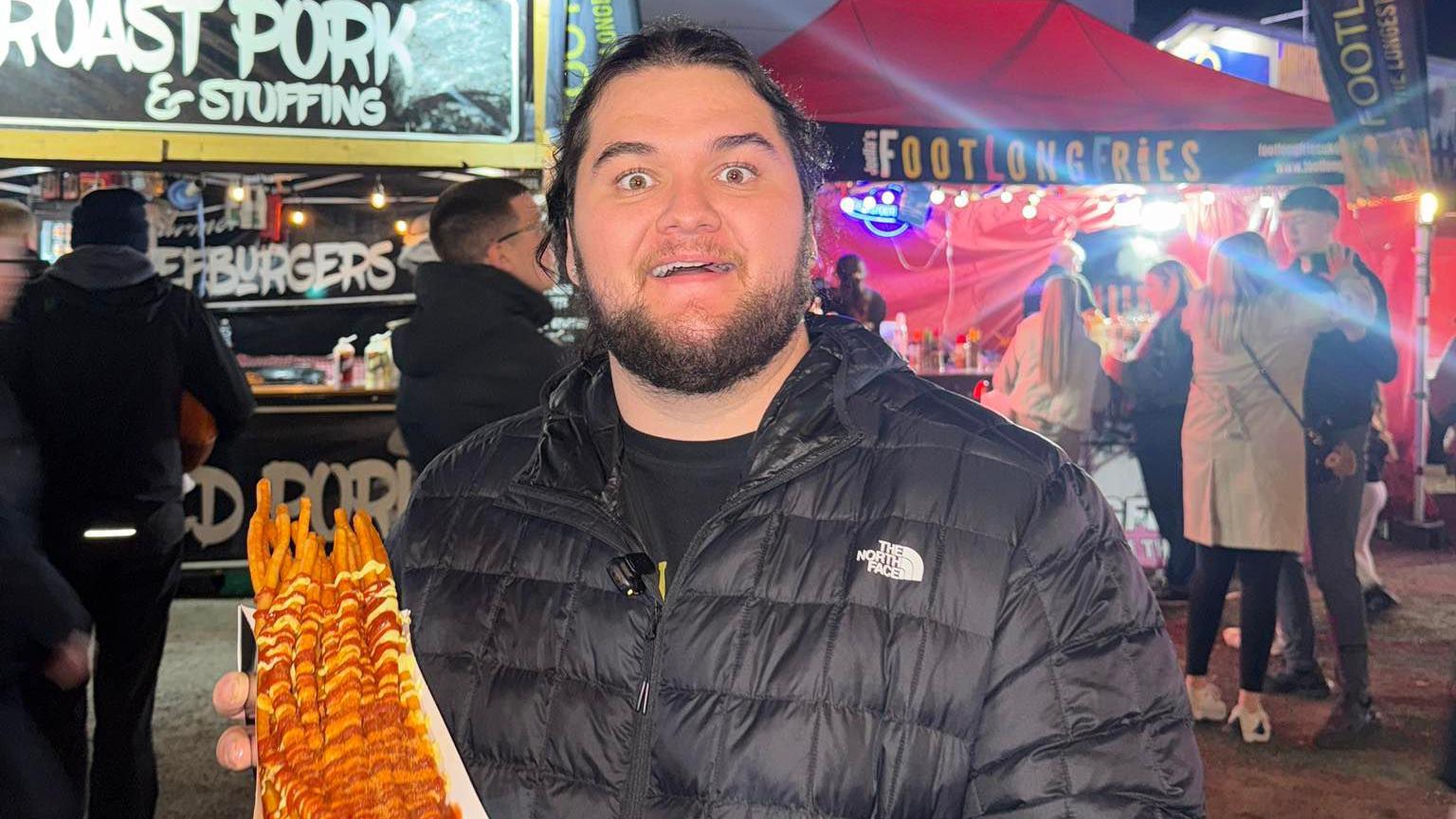 A man with brown hair tied back and a short brown beard is pulling a bewildered face and looking into the camera whilst holding a tray full of footlong fries with white and red sauce on it. He is wearing a North Face black coat and food stalls can be seen behind him.