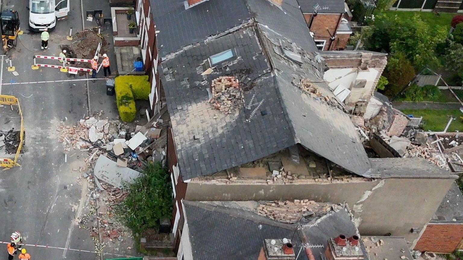 An aerial photo of a destroyed house following an explosion. Workers wearing hi vis are visible as well as a white van.
