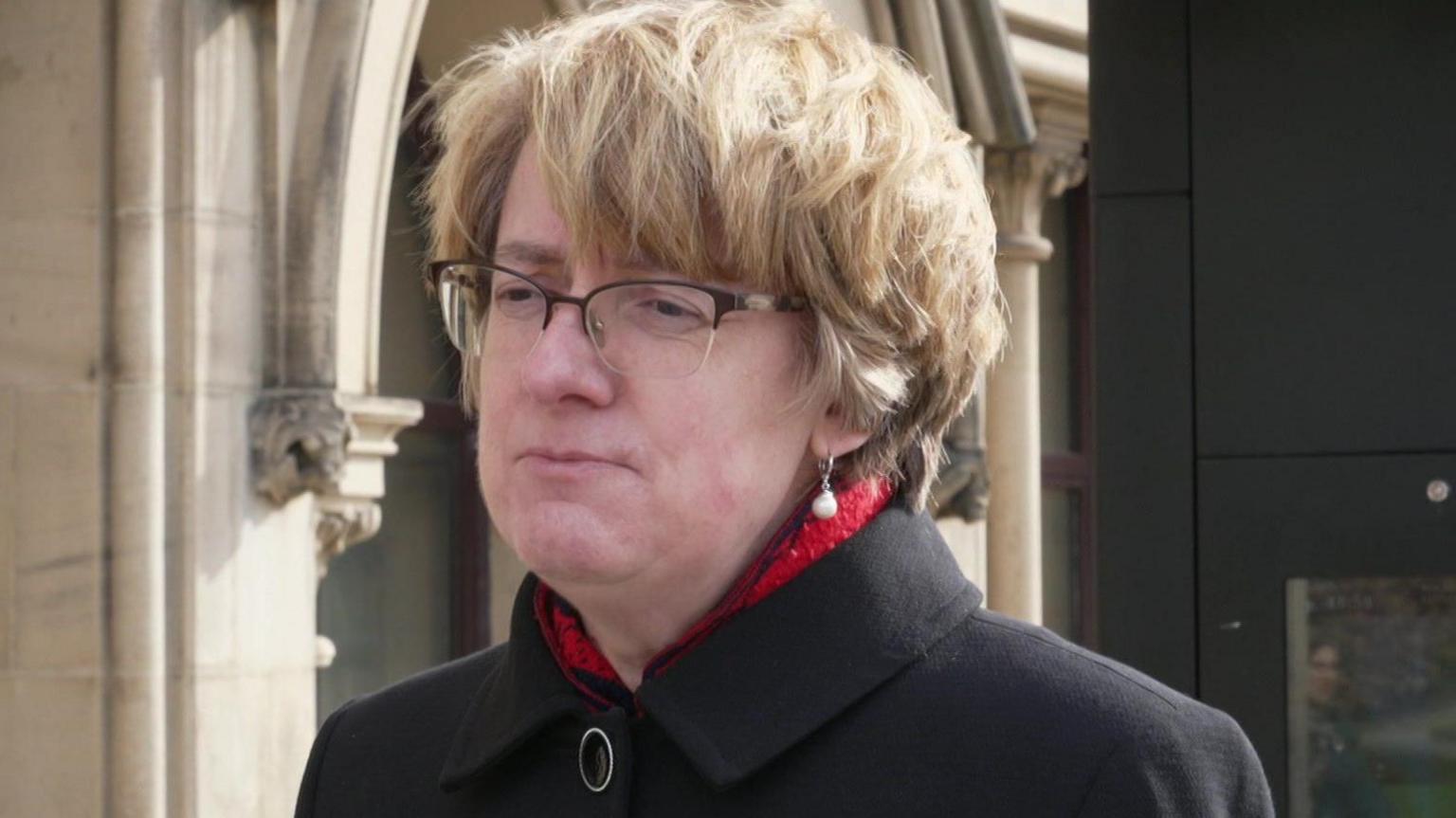 A woman with short, brown hair and glasses. She wears a black coat and a red silk scarf.