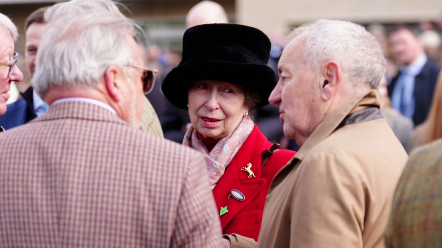 Princess Anne is in the centre of the photo. She is wearing a read coat covered in broaches. She has pearl earrings, a pink scarf and a fluffy black hat.