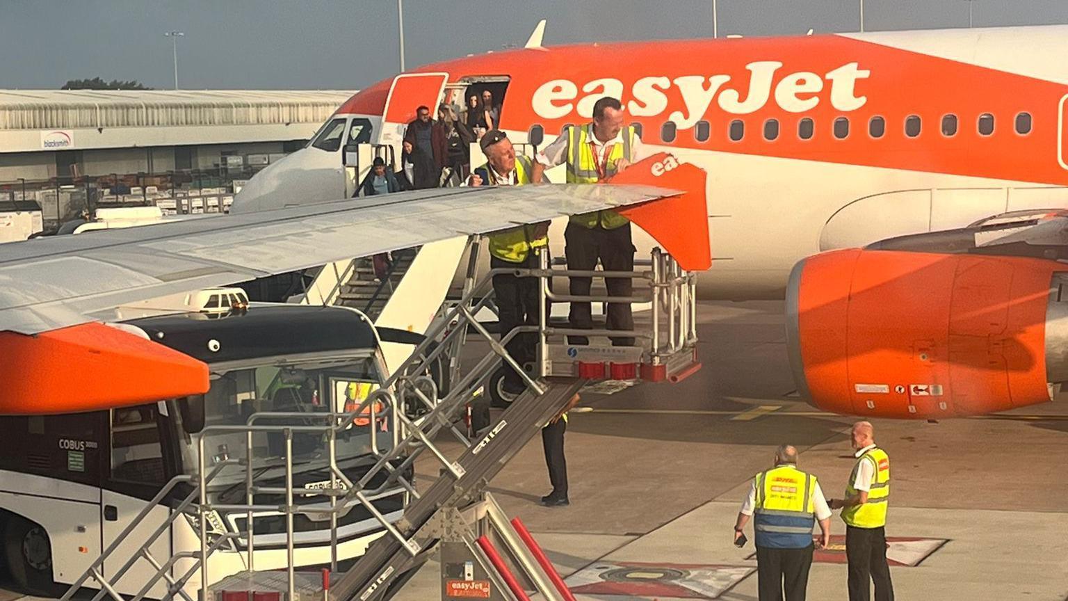 Two airport workers in yellow high-visability jackets can seen standing on a cherry picker inspecting the tip of a wing that's clipped off a plane. Passengers are aslo seen going down the stairs from the other EasyJet plane as two other safety workers stand onthe airfield.