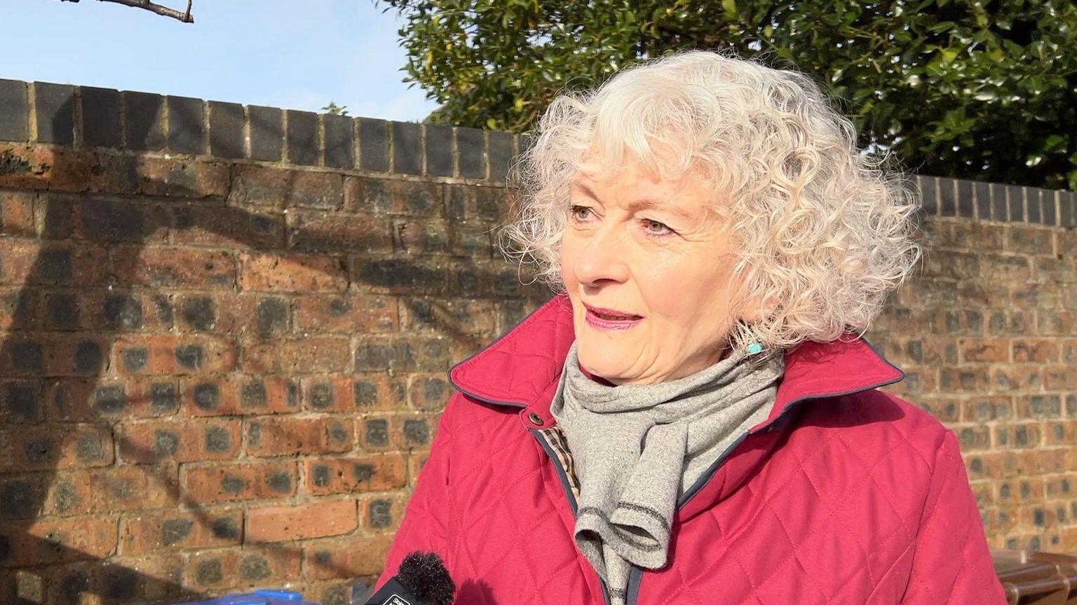 A woman who has white hair and is wearing a red coat stands on a driveway in the sunlight. 