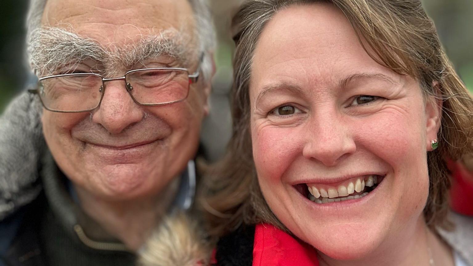 Gill, who has short brown hair, takes a selfie with her father, who is clean shaven with glasses, short white hair and thick eyebrows. Both are smiling warmly at the camera.