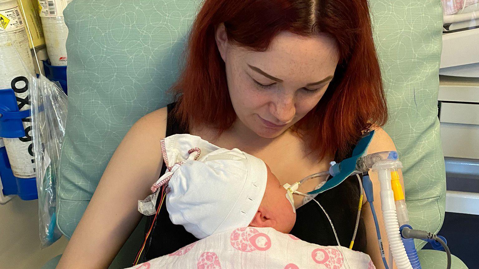 Tia with dark red hair is sitting in a green soft chair wearing a black sleeveless top, looking down at the baby cradled against her chest. The back of Arabella's head, wearing a white hat is visible as is part of the white and pink blanket that covers her. Tubes can be seen coming from her face. Oxygen cannisters are visible in the background.