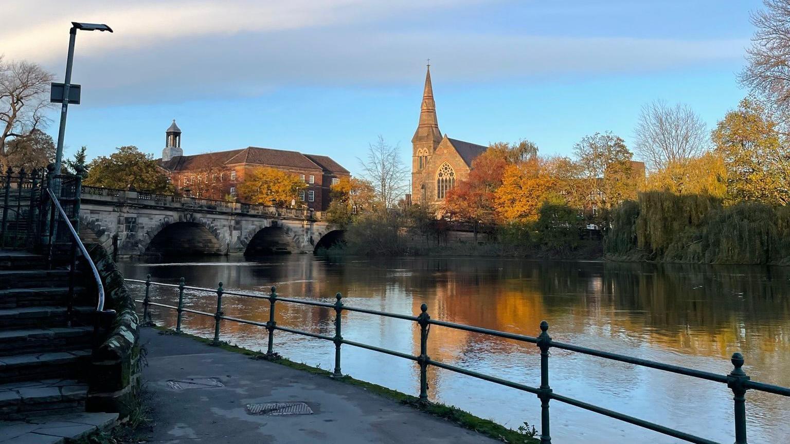 A picture of the River Severn in Shrewsbury. The sky is blue with white clouds, and you can see a bridge that goes over the river. There is a church in the background, with green, orange and yellow trees that are partially bathed in sunlight