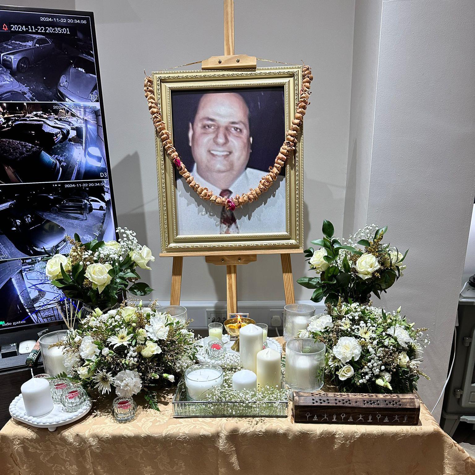 A picture of Mr Verma with a flower garland draped across it. There are white candles and flowers on a table beneath his photograph.