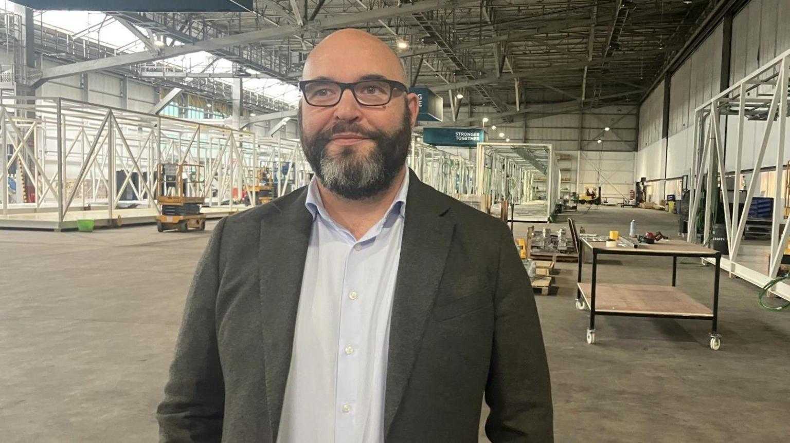 Michael has a full beard, black with patches of grey hair around his chin. Suited and wearing dark rimmed spectacles, he is standing in an air hanger.
