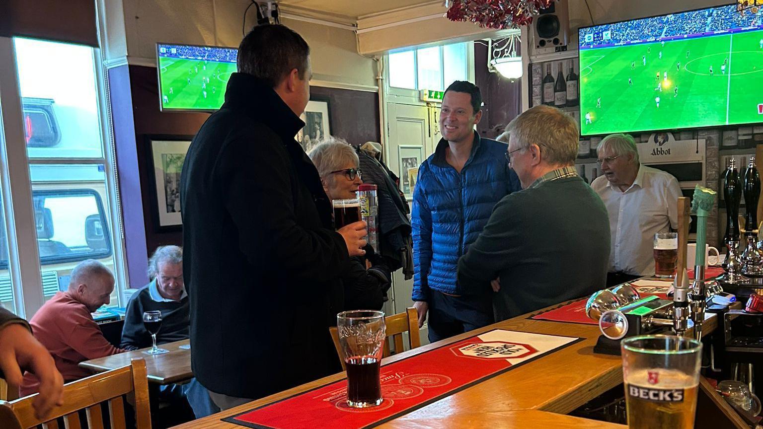 A group of people gather around a bar in a traditional British pub. There is a sports game on the TV, and people can be seen sat around a table in the background.