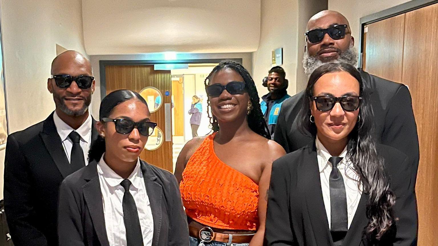 A woman in orange top and jeans in dark glasses, posing in a corridor with two women and two men in dark suits, black ties, white shirts and dark glasses.