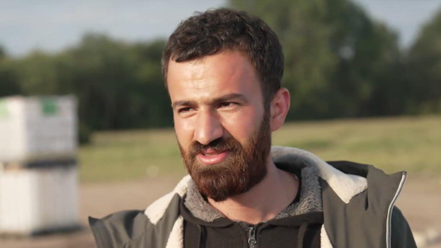 A man with a thick brown beard wearing a black jacket and coat. He is standing in front of a green field.