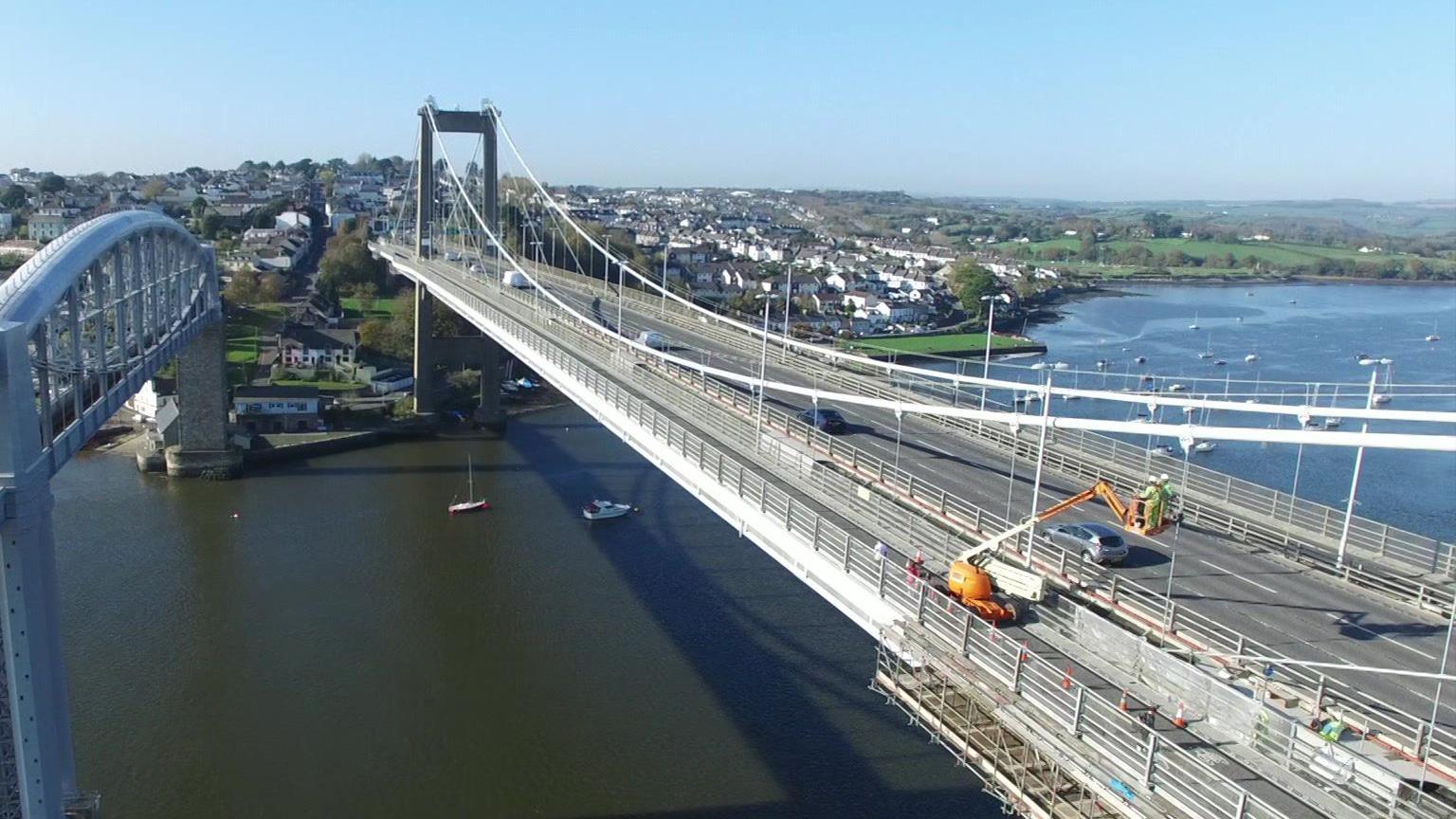 An aerial view of a large suspension bridge, with a river running beneath it. There is an orange cherry picker at the side of the bridge, with a couple of people working at height in it.