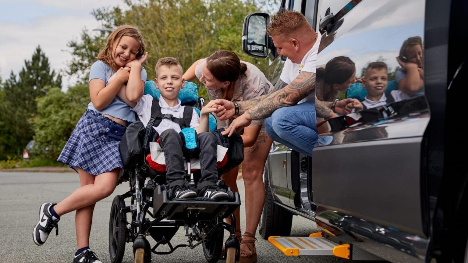 A family of four are stood around a mini-van. On the left is a girl who is holding hands with a boy who is in a wheelchair. The boy's parents are to the right and looking towards him