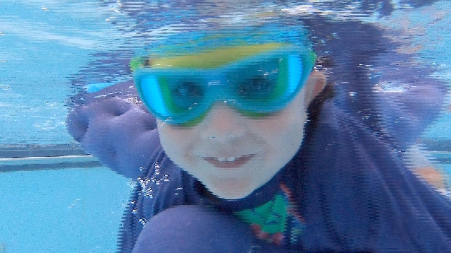 A toddler is underwater in a swimming pool smiling. They are wearing a pair of swimming goggles and a cape is billowing behind them.
