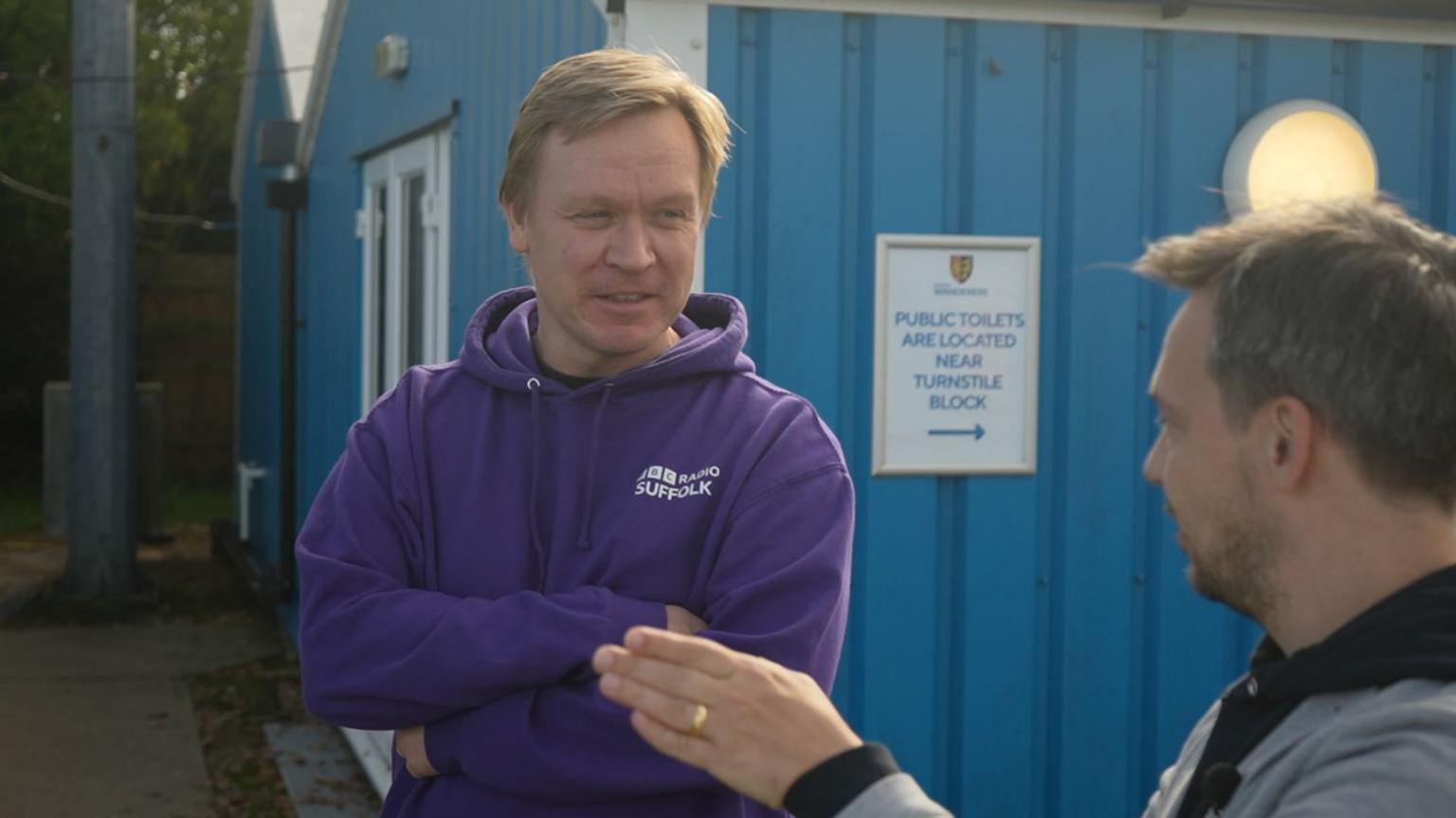 Graeme McLoughlin smiles at Jacques, as the pair catch up. McLoughlin is wearing a purple BBC Radio Suffolk-branded hoodie. 