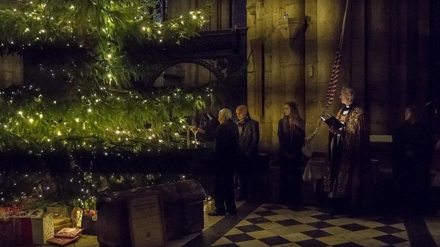 Jackie McCowliff hanging the miner's safety lamp on the tree. He has white hair and is standing next to another man, Sally Lockey who has long brown hair and the Reverend Canon.