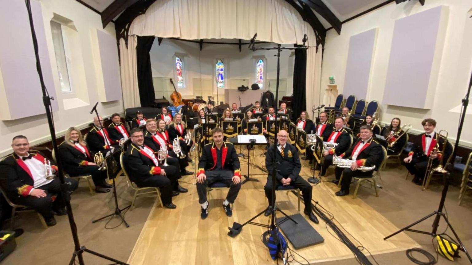 Dozens of brass band members are sitting in a U-shape with their instruments and smiling at the camera. Sam Fender, also in the red and navy band uniform, sits at the front alongside Peter Lawson.