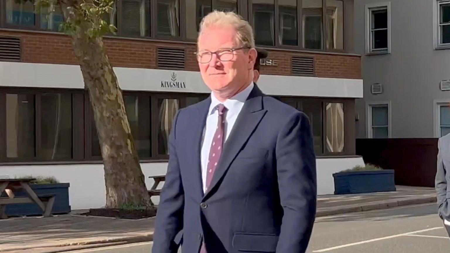 Andrew McLaughlin, the chief executive of the Government of Jersey, walks along a street in St Helier on a sunny day while wearing a navy blue suit, white shirt and maroon tie. He is wearing glasses and has trimmed blond hair. An office building for a firm called Kingsman is on the other side of the road along with a tree, two benches and two planters.