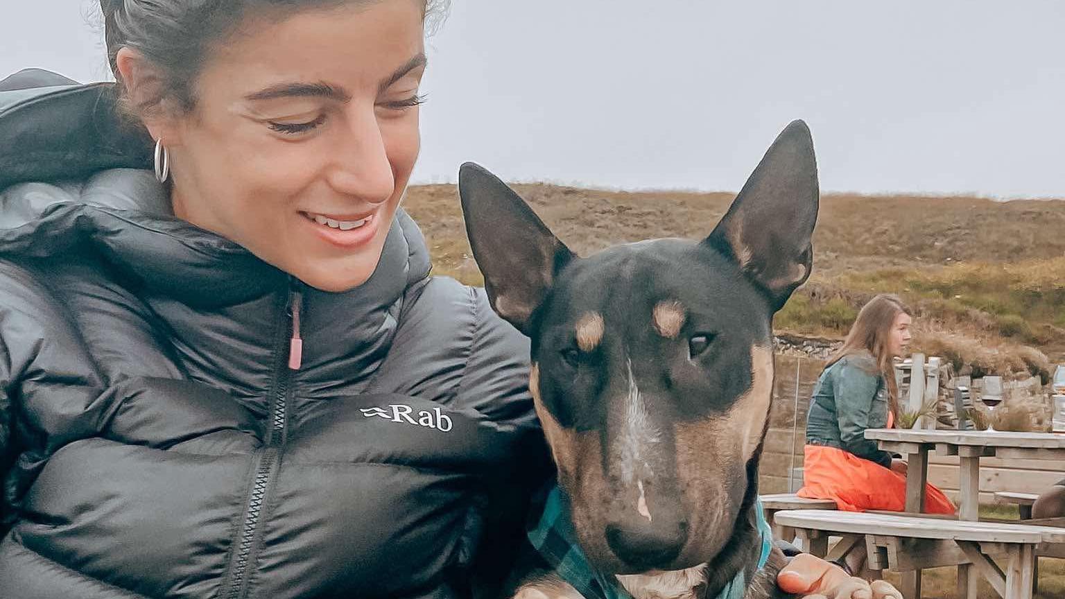 A woman wearing a black puffer jacket with black hair looks down at a dog with brown and tan markings.