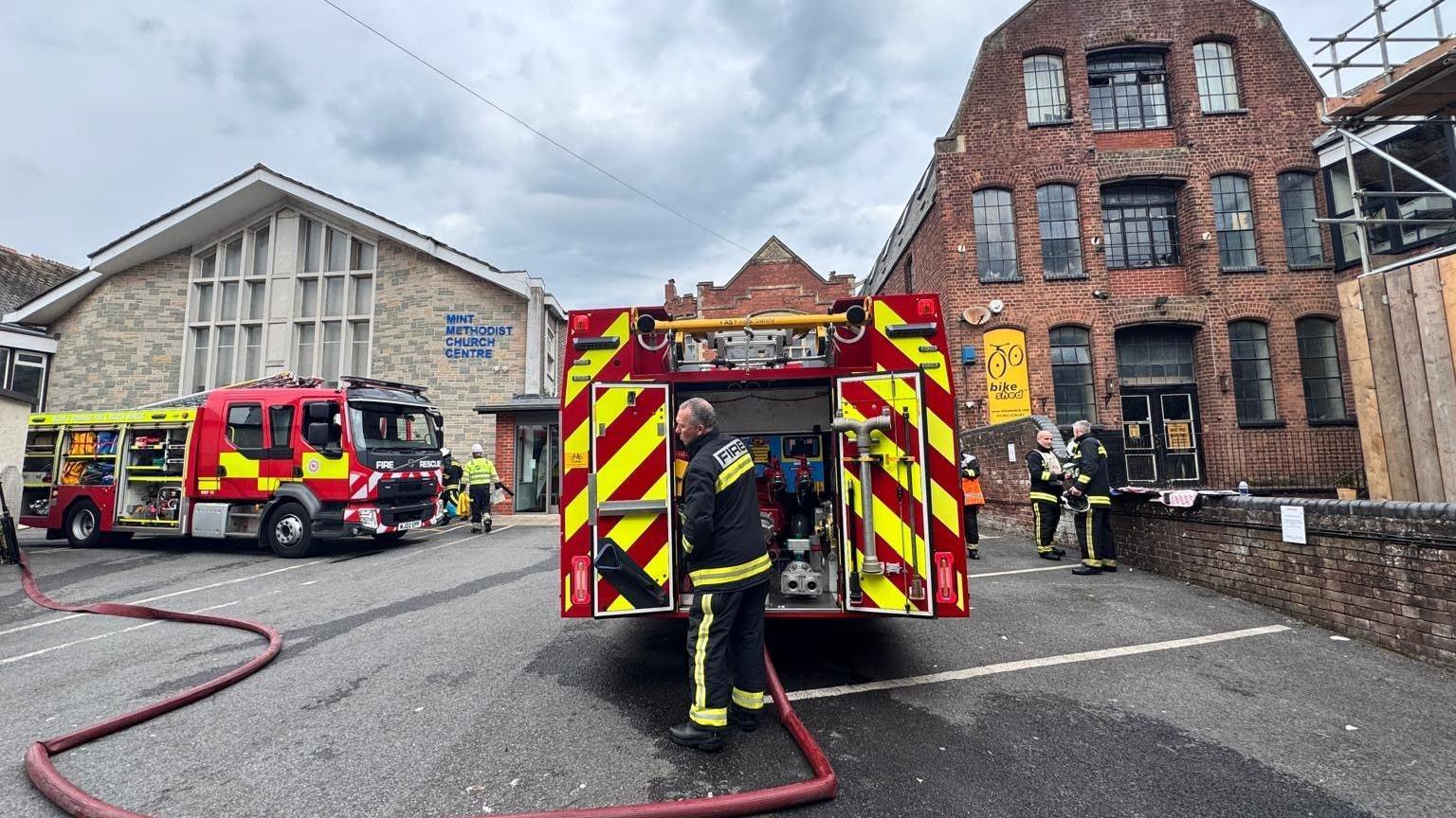 Fire engines and firefighters at the scene of a substation fire in Exeter. There are hose reels on the ground and fire crews dotted around in the background.
