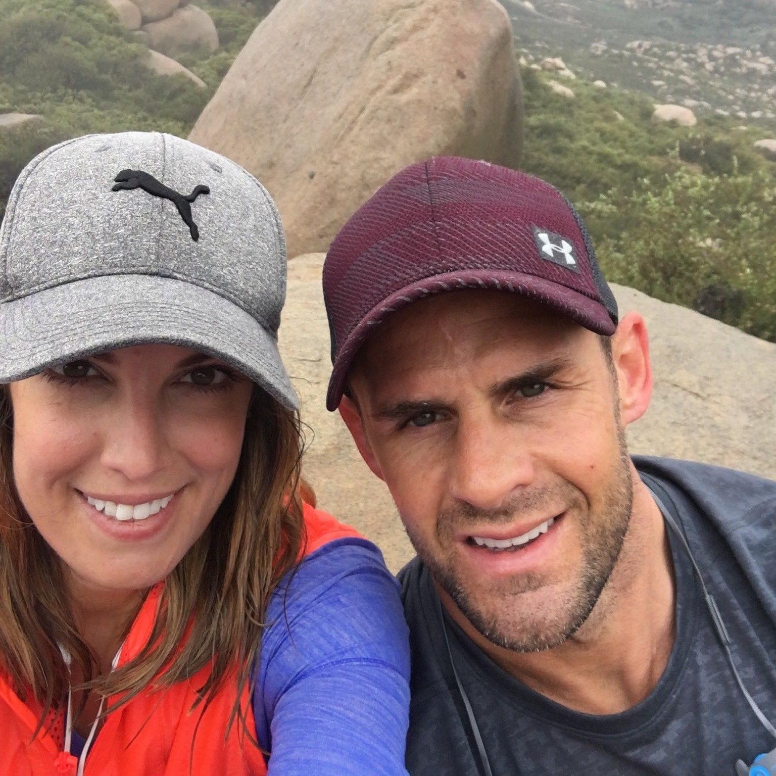 Andrea, wearing a grey baseball cap while hiking, smiles for a selfie next to husband Lee, who is wearing a maroon baseball cap